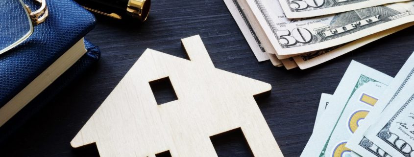 Wooden house on table with hundred dollar bills money and journal pen and glasses