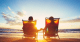 retired couple sitting on the beach in lounge chairs together watching the sunset